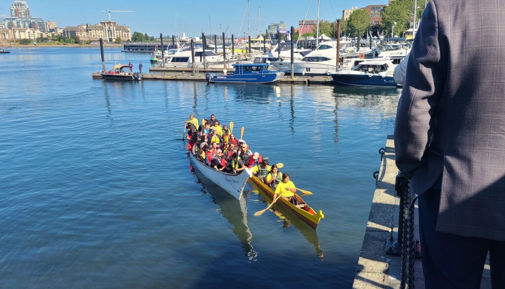 Two canoes are filled with Canadian senators, MPs, former chiefs, and other dignitaries dressed in casual clothes and wearing life vests. They are being greeted by a representative of Songhees Nation on the first day of Victoria Forum 2022 while a group of other conference attendees wait on the embankment above the canoes.