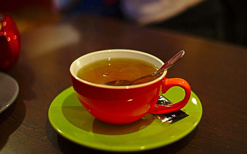A red teacup with tea and a spoon inside rests on a lime green saucer, which itself rests on a warm-wooden tabletop.