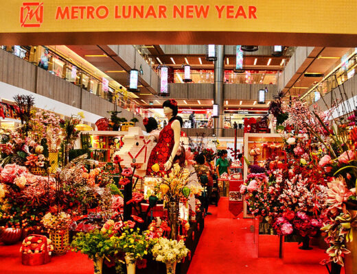 A storefront is jam-packed with displays of fresh flowers in containers of all shapes and sizes. At centre is a black-haired manikin shaped like a woman wearing a red sleeveless dress. The banner at the top of the image says "Metro New Year".