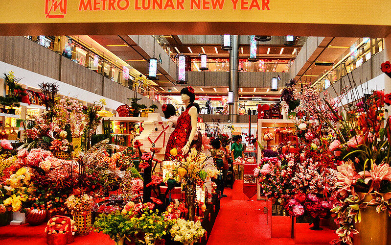 A storefront is jam-packed with displays of fresh flowers in containers of all shapes and sizes. At centre is a black-haired manikin shaped like a woman wearing a red sleeveless dress. The banner at the top of the image says "Metro New Year".