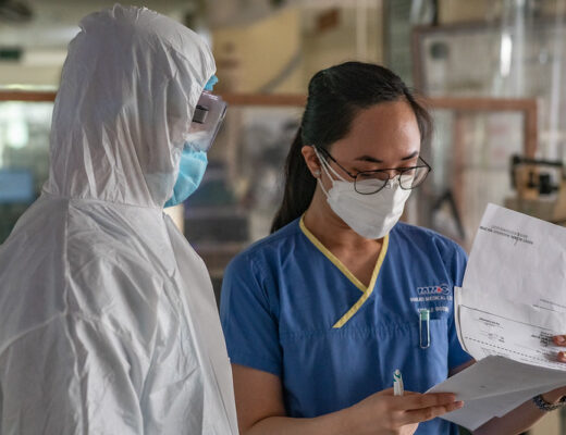Two healthcare professionals care for a patient in the Philippines. One wears a full PPE suit, the other is in blue scrubs. Both Filipino people are wearing face masks.