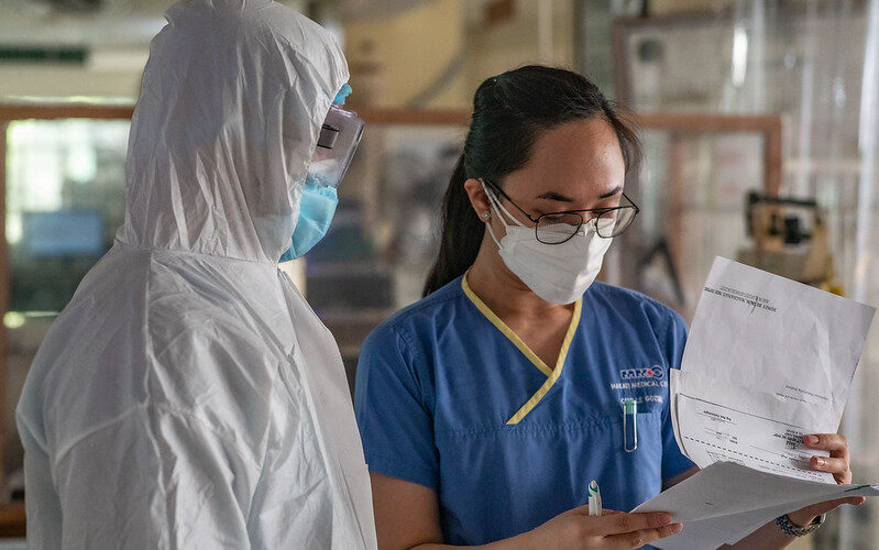 Two healthcare professionals care for a patient in the Philippines. One wears a full PPE suit, the other is in blue scrubs. Both Filipino people are wearing face masks.