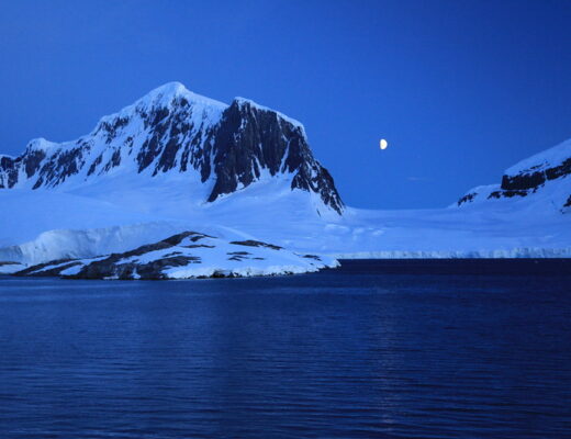 A waning gibbous moon hovers over the nighttime horizon, flanked by snow and ice-covered mountains in Antarctica.