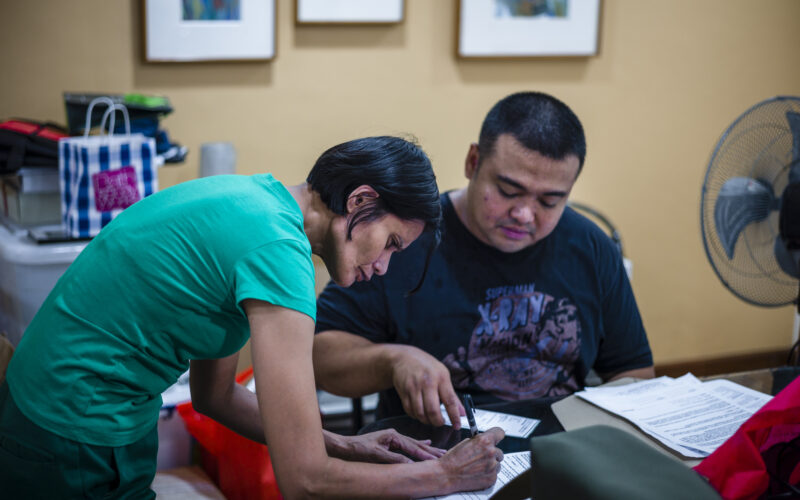 A Filipina domestic worker with short hair bends over a desk and she is writing something. A Filipino man is seated at the table she's bending over and pointing out a detail on an smartphone. The woman, Emelyn, was taught how to read, write, and use the computer by her employers.